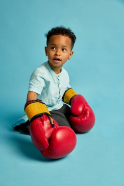 Mutlu çocuk mavi arka planda oturan boks eldivenleri giyer. Güç ve mizah gösterir. Çocukluk, spor, ifade, aile, eğlence, duygu, oyun, güç, güzellik, neşe kavramı.