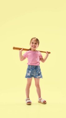 Omuzlarının arkasında tahta sopa tutan gülümseyen küçük kız rahat duruyor ve sarı arka plana karşı kendine güvenini gösteriyor. Çocukluk, yaz, spor, oyun, enerji, sağlık kavramı.