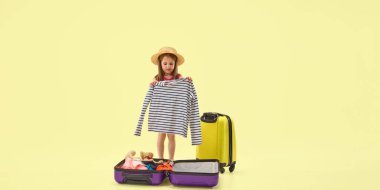 Yazlık şapkalı küçük kız Pastel stüdyo arka planına karşı giysiler ve oyuncaklarla bavul hazırlıyor seyahat macerasına hazırlanıyor. Çocukluk kavramı, tatil, gezi, yaz, neşe, planlama.
