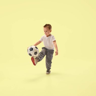 Odaklanmış küçük çocuk, pastel sarısı stüdyo arka planına bakarken futbol topunu ayaklarıyla havada kontrol ediyor. Tatil kavramı, çocukluk, yaz, spor, sağlık, disiplin.