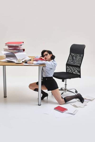 Портрет молодой женщины, выглядящей измученной лежащей головой под перегруженным столом в офисной среде на белом фоне. Концепция офисного образа жизни, человеческих эмоций, социальных проблем, бизнеса