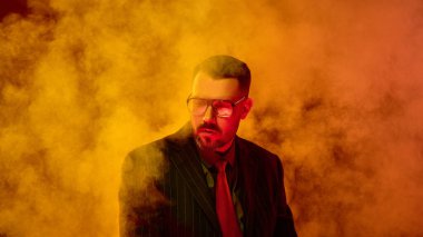 Çizgili takım elbiseli bir adam, dramatik ışık altında turuncu dumanların arasından düşünceli ve yorgun bir ifadeyle aşağıya bakıyor. Duygusal izolasyonu ifade ediyor. Kriz kavramı, depresyon, psikoloji, stres.