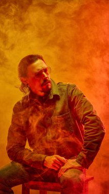 Gözlüklü adam kırmızı-turuncu neon ışıkta oturuyor. Gergin vücut duruşu direnç ve sessiz öfke gösteriyor. Duygu kavramı, erkeklik, depresyon, psikoloji, tükenmişlik.