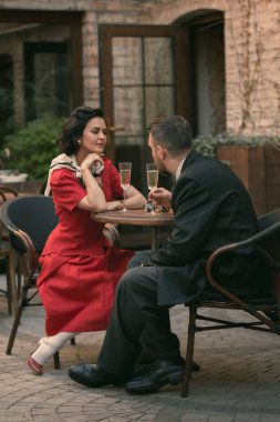 Geçmişten giyinmiş bir çift yuvarlak masaya oturup, içkilerini yudumluyor, hoş bakışlarını ve klasik sessizliğini paylaşıyorlar. Sahne sakin romantizmi gösteriyor. Gizlenmek için uygun. Bağbozumu, aşk, nostalji kavramı.