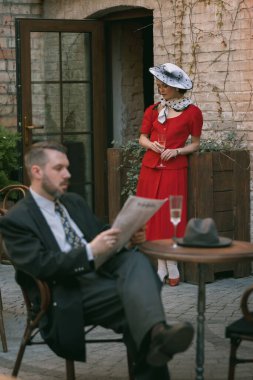 Zarif, şık, kırmızı bir elbise giymiş güzel genç bir kadın elinde içki olan ahşap ekicinin yanında duruyor ve açık havada kitap okuyan adama bakıyor. Eski moda, aşk, nostalji, romantik randevu kavramı..