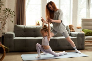 Yetişkin bir çocuk, yoga seansında etkileyici bir şekilde bacak esnetme hareketine yardımcı olur, paspasın üzerinde çocuk dengeleri, rahat oturma odasındaki hareketlere odaklanır. Evde ebeveynlik mesleği kavramı. Ad