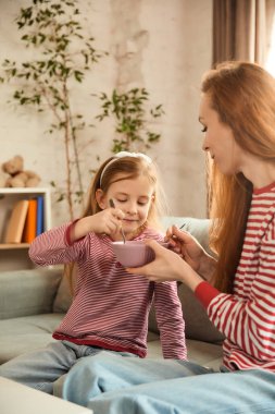 Çocuk mutlu bir şekilde pembe kaseden yetişkinlere yemek yiyor, modern oturma odasındaki gri kanepede otururken birlikte yemek yiyorlar. Sağlıklı yaşam tarzı, ebeveynlik, öğrenme kavramı. ad