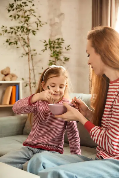 Çocuk mutlu bir şekilde pembe kaseden yetişkinlere yemek yiyor, modern oturma odasındaki gri kanepede otururken birlikte yemek yiyorlar. Sağlıklı yaşam tarzı, ebeveynlik, öğrenme kavramı. ad