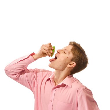 Pembe gömlekli genç adam portresi başını arkaya eğiyor, ekşi yeşil limonu beyaz stüdyo arka planında tadıyor. Doğal gıda, organik beslenme, diyet, vitamin kavramı. Ad