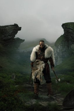 Bileşik kolaj. Kürk pelerinli vahşi Viking sisli tepeler ve vahşi araziler tarafından çerçevelenmiş baltalarla dolu uçurumlar arasında duruyor. İskandinav, doğa, liderlik, tarih kavramı