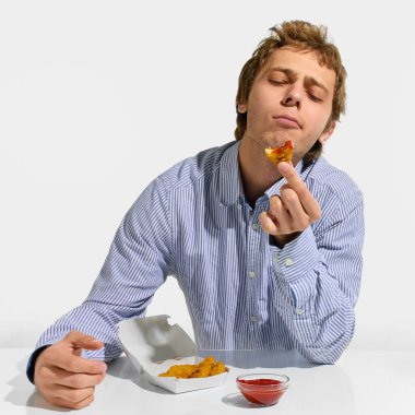 Elinde ketçaplı tavuk külçesi tutan ve gözleri zevkten kapalı bir adam. Masadaki zengin lezzeti beyaz stüdyo arka planında takdir ediyor. Geleneksel, fast food, yemek servisi, paket servis.