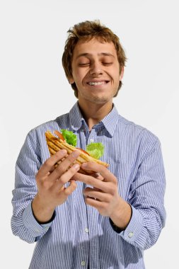 Salata ve domatesle dolu ızgara sandviçi nazikçe tutarken gözleri kapalı gülümseyen bir adam beyaz arka planda tattan zevk alıyor. Yemek tatmini kavramı, sokak yemekleri.