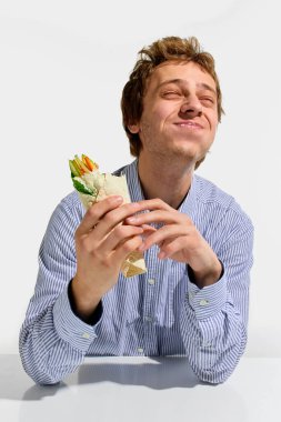 Neşeli bir adam, sebzeli dürüm çiğniyor, gözleri kapalı ve çizgili gömlekle gülümsüyor, elleri beyaz arka planda tortilla tutuyor. Geleneksel, yemek servisi, fast food, beslenme, paket servis.