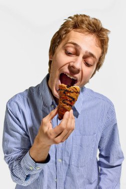 Çizgili gömlekli bir adam beyaz arka planda neşeli bir pozda çikolata jöleli gevrek kruvasan yemek için ağzını açıyor. Fast food, beslenme, geleneksel, paket servis, yemek.