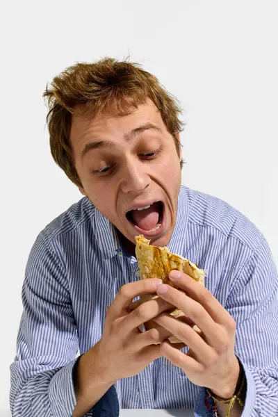 Genç adam ızgara shawarma böreği tutarken hevesle ağzını açıyor, beyaz arka planda ısırmadan önce neşeli bir tepki gösteriyor. Shawarma kavramı, yemek, iştah, neşe, sokak yemekleri