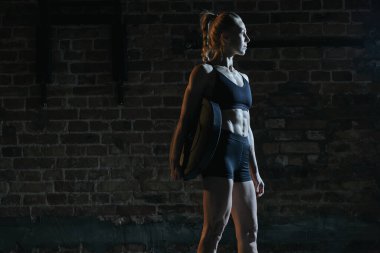Siyah spor kıyafetli bayan sporcu dramatik bir spor salonunda büyük yuvarlak bir plaka tutuyor ve önüne bakıyor. Güç kavramı, beden eğitimi, sağlıklı vücut, odaklanma.