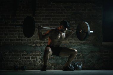 Gömleksiz, kaslı bir adam, yoğun bir şekilde, dramatik spor salonu ışıklandırması altında omuzlarında barla derin halterler üzerinde çömeliyor. Spor salonu kültürü, güç, direnç, eğitim, sağlıklı güç kavramı