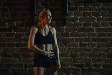 Spor salonunda rahat duran atletik kadın egzersizden sonra karın kasları üzerinde dinlenerek güvenle gülümsüyor. Formunu ve gururunu tuğla duvara karşı sergiliyor. Spor anlayışı, aktif ve sağlıklı yaşam tarzı