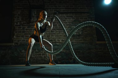 Savaş halatlarıyla çalışan kadın sporcu çömelme pozisyonunda simetrik dalgalar yaratarak patlayıcı enerji ve kontrol gösteriyor. Spor anlayışı, aktif ve sağlıklı yaşam tarzı, vücut geliştirme