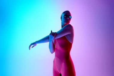 Kadın yüzücü, özgüvenle kırmızı takım elbise ve gözlüklerle gradyan stüdyo arka planına karşı duruyor. Hazırlık, odak ve iç gücü tasvir ediyor. Motivasyon, spor, disiplin, kararlılık kavramı