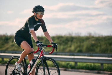 Kırsal yolda bisiklet süren, kask takan ve güneş gözlüğü takan, gün ışığında düz yolda ilerleyen kadın bisikletçinin yan görüntüsü. Spor, spor, rekabet kavramı.