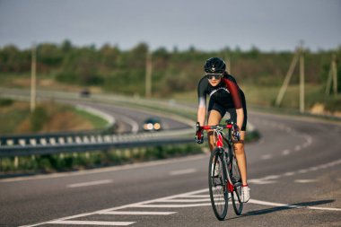Açık yolda yokuş yukarı giden kararlı kadın bisikletçinin dinamik fotoğrafı pedalları yoğun odakta, kask takıyor ve dar bir forma giyiyor. Ekstrem spor kavramı, şampiyonluk, çaba.