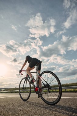 Bisikletçinin gün batımına doğru sürüşünün alçak açılı fotoğrafı, asfalt boyunca açık ufukta uzun gölgeler bırakıyor. Mesafe kavramı, çaba, ekstrem spor, yalnızlık, güç, kararlılık.
