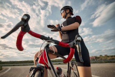 Düşük açılı kadın bisikletçinin yol kenarındaki eğri, bisiklet iskeleti ve gidonu ön planda dururken eldiven ayarlarken görüntüsü. Ayrıntılı fotoğraf. Spor eğitimi kavramı, duraklama, vites, hazırlık