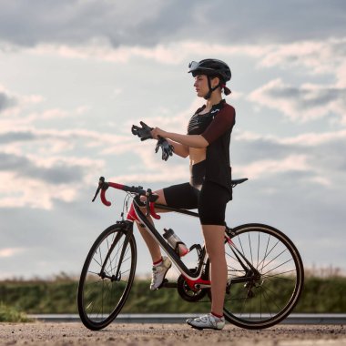 Kırmızı ve siyah yol bisikletinin yanında duran formda bir kadın, bulutlu gökyüzünün altında eldivenlerini takıyor, antrenmandan önce hazırlanıyor. Yoğun spor eğitimi kavramı, açık hava fitness, vites