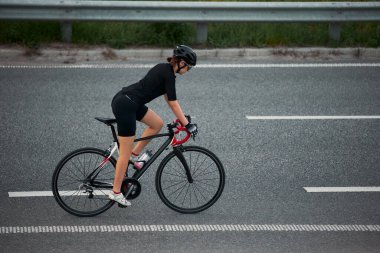 Yan görüntü fotoğrafı. Otoyoldaki bariyerlerin önünden geçen kadın bisikletçi antrenmanlara odaklanmış. Hareket bulanıklığı etkisi. Spor eğitimi kavramı, disiplin, bisiklet çabası, odaklanma
