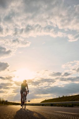Gün batımında, dramatik gökyüzü ve güçlü ışık patlaması ile çerçevelenmiş bir şekilde güneş ışınlarına doğru bisiklet süren bir kadın. Maraton. Ekstrem spor, özgürlük, berraklık, eğlence, enerji kavramı