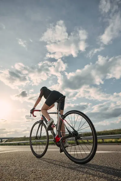 Bisikletçinin gün batımına doğru sürüşünün alçak açılı fotoğrafı, asfalt boyunca açık ufukta uzun gölgeler bırakıyor. Mesafe kavramı, çaba, ekstrem spor, yalnızlık, güç, kararlılık.