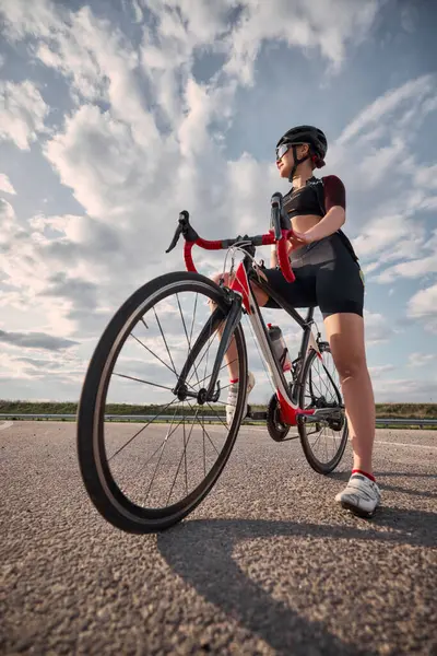 Düşük açılı fotoğraf, yarış bisikletinin yanında duran bir ayak ileri, mesafeye bakan, arka planda yumuşak bulutlarla dolu bir kadın. Spor eğitimi kavramı, vizyon, hırs, dinlenme.