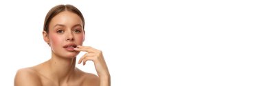Çekici genç bir kadın, elleri dudaklarına yakın, omuzları beyaz arka planda, çıplak bir şekilde ileriye bakıyor. Doğal tırnaklar ve nazik makyaj. Cilt bakımı, doğal görünüş, zarafet, yüz güzelliği.