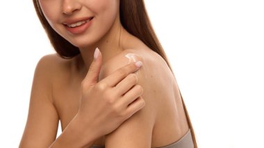 Kadın elinin omuz derisine beyaz nemlendirici krem sürerken, gülümseyen yüz hafif görünür, yumuşak ışık ve sıcak ton cilt efektini arttırır. Vücut bakımı kavramı, hidrasyon rutini