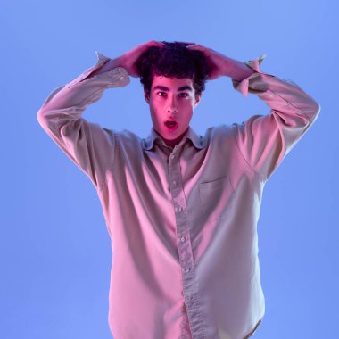 Mavi stüdyo arka planına karşı yumuşak pembe-mavi ışıkta elleri kafasında poz veren, aşırı büyük tişörtlü çocuk. Sürpriz kavramı, karmaşa, ifade,