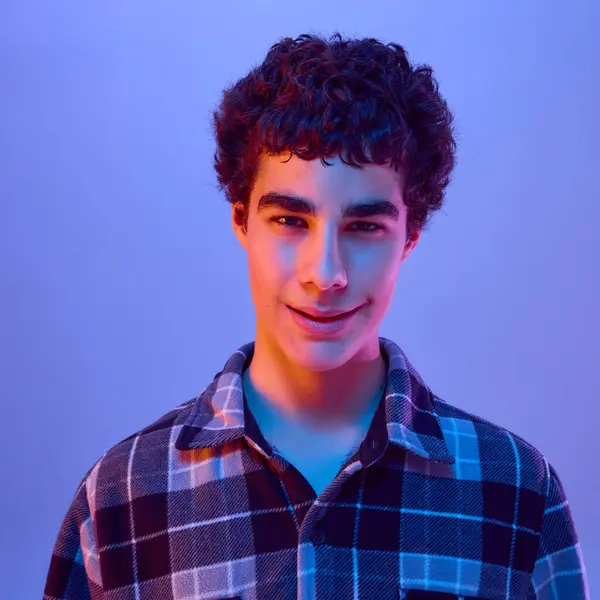 Kaş kaldıran ve gevşemiş bir ifadeyle gülümseyen, neon ışığı altında arka planda işaretli gömlek giyen genç bir çocuğun yakın çekimi. Güzellik kavramı, refah, rahatlama, merak.