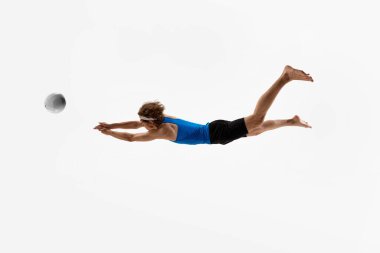 Voleybol oyuncusu, şık üniformalı, elleri havada topa uzanmak için öne doğru dalıyor, sıçrarken, beyaz stüdyo arka planına karşı tüm dikkatini gösteriyor. Spor anlayışı, tepki