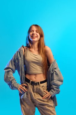 Genç kız neon ışıkta mavi arka planda günlük mahsul bluzu ve açık kot gömlek giyerek neşeli hareketlerle kahkahalar atıyor. Mutluluk kavramı, insan duyguları, moda, gençlik