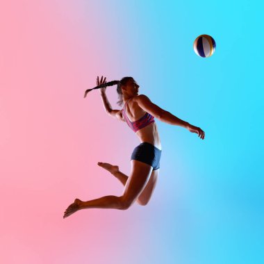 Atletik kadın, şort ve top giymiş, voleybol oynamak için atlayan pembe-mavi stüdyo arkaplanına karşı. Voleybol yeteneği, güç ve yüksek enerjili spor performansı kavramı.