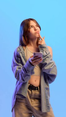 Genç kız, mavi arka planda akıllı telefonu elinde tutarak dudağına dokunarak karar verme sürecini ifade ediyor. Merakın, şaşkınlığın, düşüncenin kavramı.