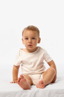 Beyaz tulum giymiş çocuk beyaz arka planda tuhaf bir ifadeyle beyaz yumuşak yatakta oturuyor ve sürünüyor. Çocukluk kavramı, masumiyet, duygu, annelik, tarz, konfor