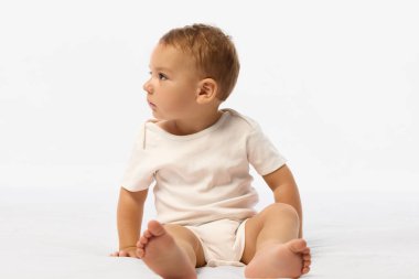 Beyaz tulumlu çocuk bacak bacak üstüne atmış ve beyaz arka planda tuhaf bir bakışla başını çeviriyor. Dikkat kavramı, keşif, büyüme, çocukluk, duygu, merak.