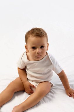 Küçük çocuk yumuşak bir yatakta oturuyor, bacakları ayrık, kaşları çatık ve beyaz arka planda ciddi bir yüzü olan bir bebek bezi tutuyor. Çocukluk kavramı, davranış, ruh hali, duygu, bağımsızlık, ifade.