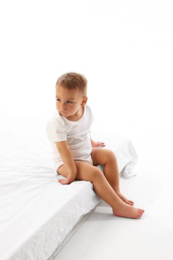 Küçük çocuk yatağın kenarında oturuyor ve eli arkada özenle aşağıya bakıyor. Beyaz stüdyo geçmişine karşı. Bağımsızlık kavramı, çocuk oyunları, çocukluk, yansıma, büyüme, geçiş.