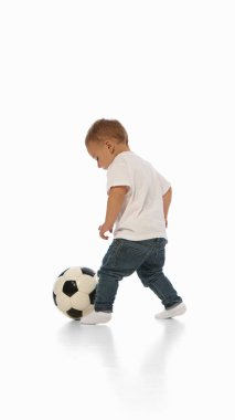 Kot pantolonlu ve tişörtlü çocuk beyaz arka planda siyah beyaz futbol topunu şen şakrak tekmeliyor. Aktif oyun kavramı, fiziksel büyüme, spor, çocukluk, motor geliştirme, merak.