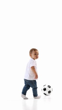 Kot pantolonlu ve tişörtlü çocuk futbol topunun yanında yürüyor, sakince omzunun üstünden beyaz stüdyo arka planına bakıyor. Hareket kavramı, bebek oyunu, spor, bağımsızlık, odaklanma, çocukluk.