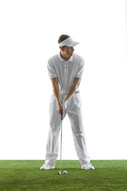 Beyazlar içinde yeşil çimlerin üzerinde duran genç golfçü sakin bir konsantrasyon ve klasik duruş ile topa vurmadan önce odaklanıyor. Golf, retro spor, odaklanma, zarafet kavramı.