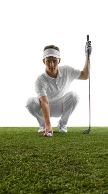 Golf kıyafeti giymiş genç adam çimenlere top koymak için çömeliyor, sürekli göz teması kuruyor ve beyaz zemine karşı mükemmel bir duruş sergiliyor. Golf kavramı, konsantrasyon, başlangıç ritüeli, hassasiyet.