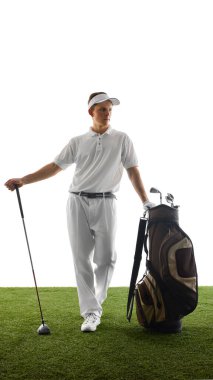 Golfçü sakince golf çantasının yanında duruyor, golf sopasını tutuyor ve beyaz arka planda golf forması giymiş bir şekilde poz veriyor. Spor aletleri, hazırlık, eğlence, klasik